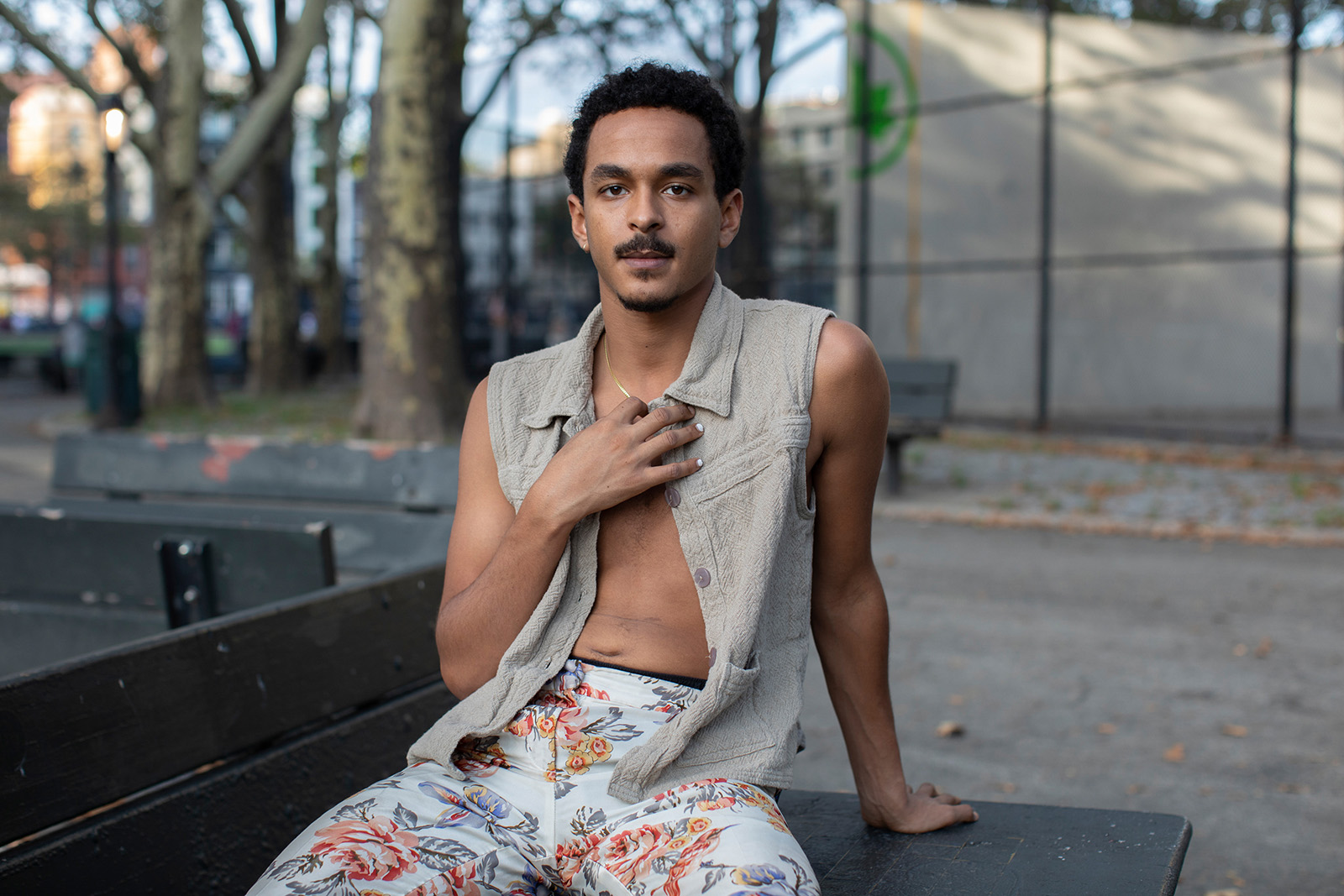 A photograph of a young man, Ahmed Mohamed, sitting on a park bench with his right hand against his chest, taken in New York in 2021