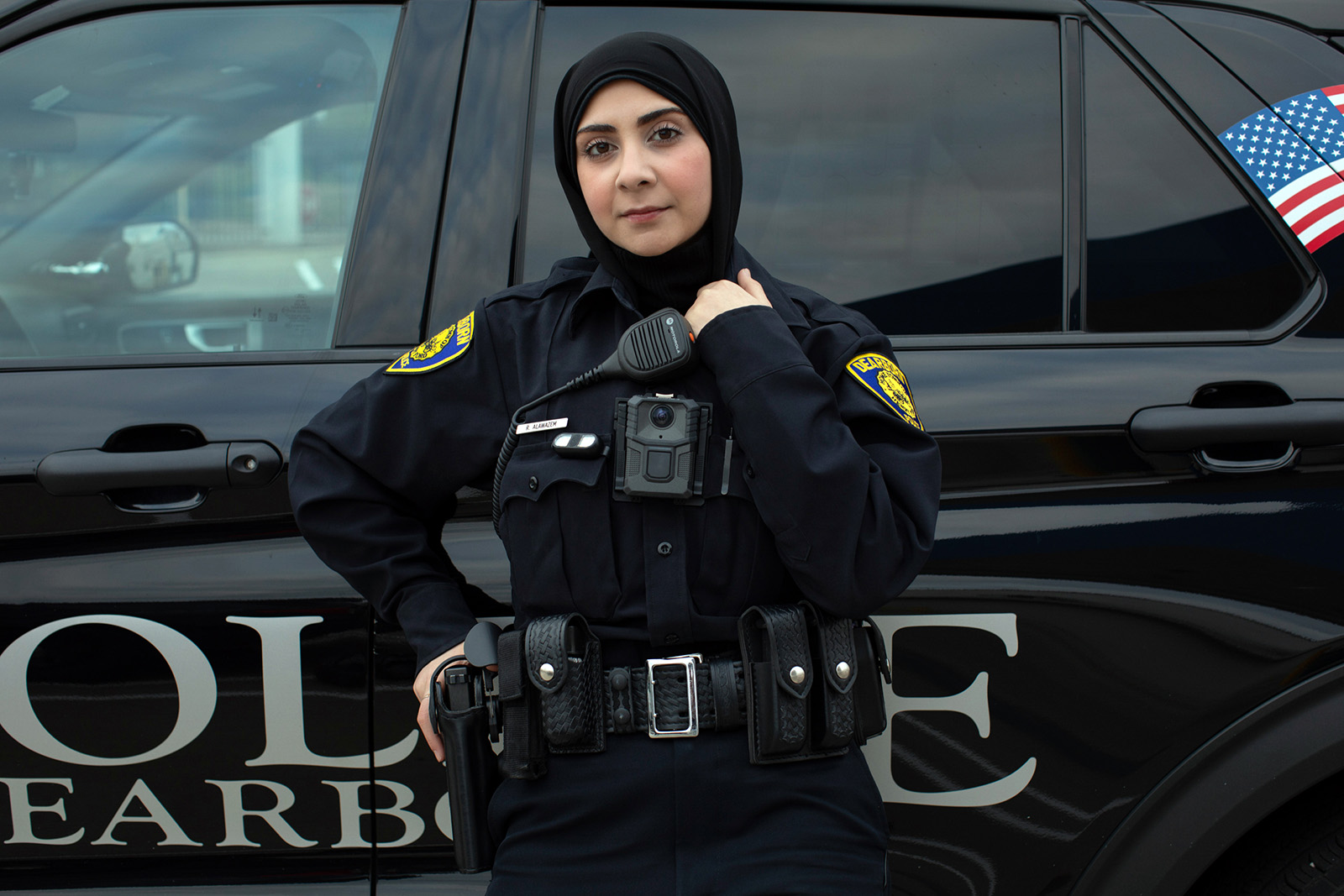 A photograph of a police officer, Raghad Alawazem, in her uniform and hijab standing in front of a police vehicle with her left hand holding her collar and right hand resting on her holstered pistol, taken in Dearborn in 2024