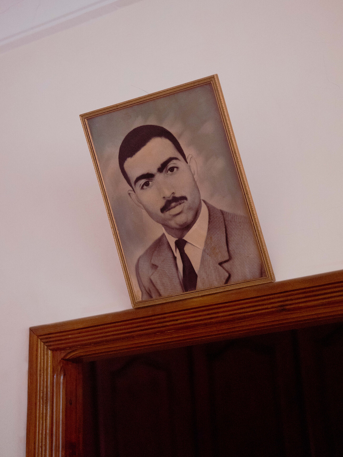 A photograph by Mayssa Jaoudat of a framed black and white head and shoulders portrait of a man in a suit and tie, hung on a white wall above a doorway