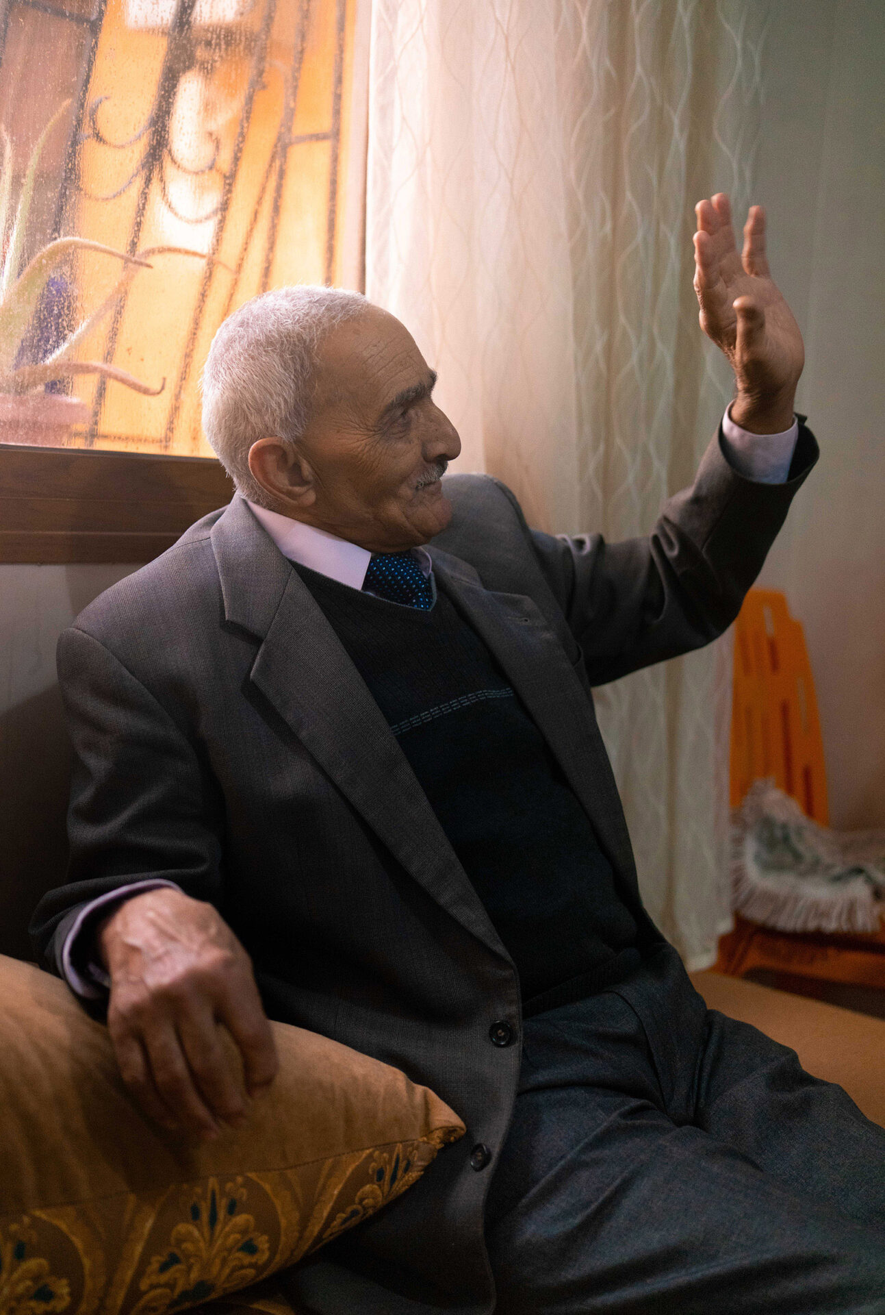 An image of an elderly man, seated indoors by a window, holding up his left arm as if greeting or saying goodbye to someone off camera. From the series Suzanne, Long Road by Mayssa Jaoudat, part of her wider photographic project Mille-Feuilles