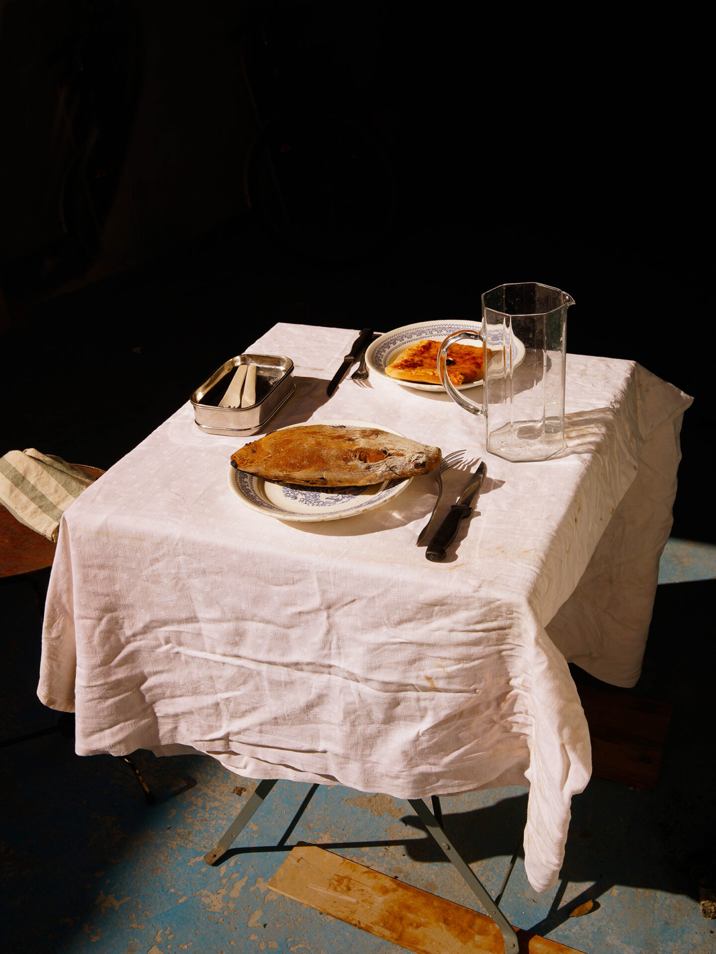 A photograph by Mayssa Jaoudat of a small table covered with a white tablecloth, on which a simple meal has been laid of two plates with bread plus an empty glass water jar and cutlery