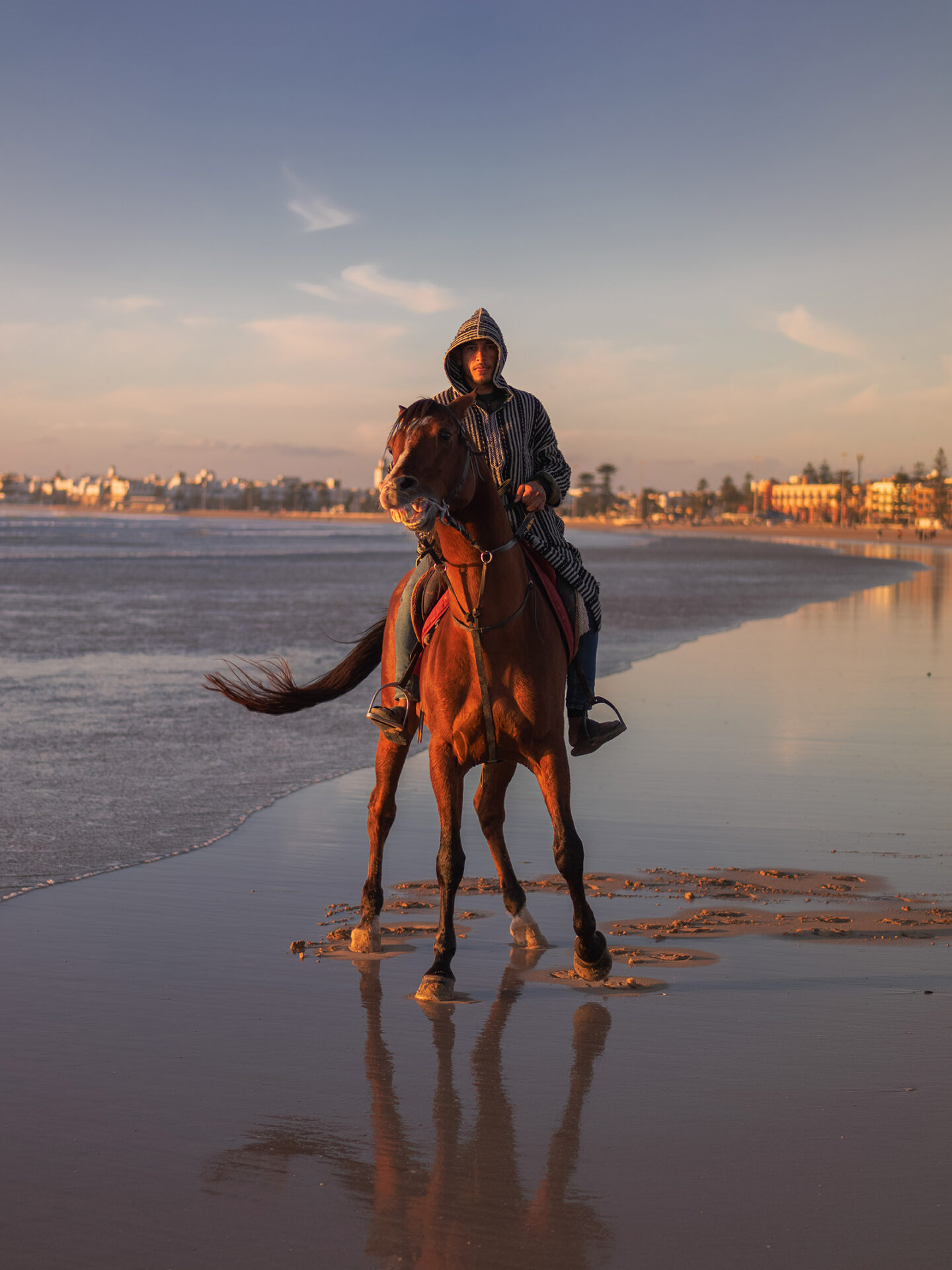 A photograph by Mayssa Jaoudat of a young man wearing a hooded jacket, on horseback at the water's edge on a sandy beach, staring directly at the camera