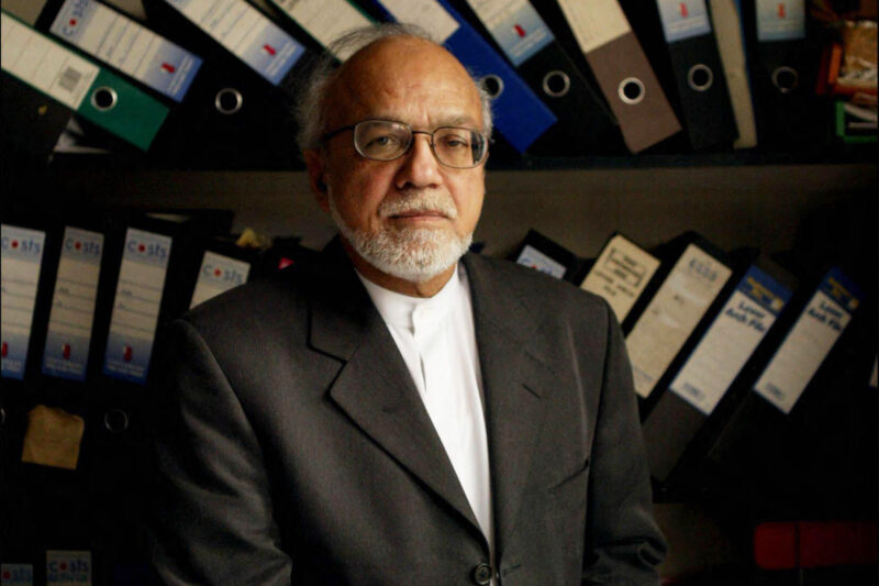 A middle-aged man in a suit and glasses. Behind him are box files leaning over onto each other, on two shelves