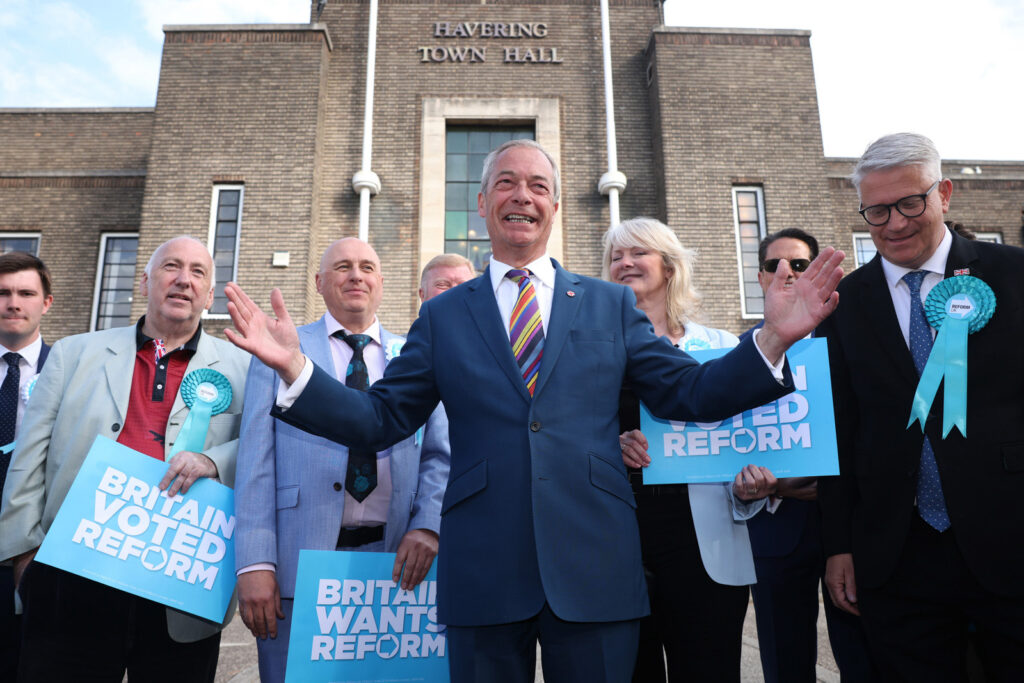 A photograph of a beaming Nigel Farage at a photocall the day after 2026 local elections, posing with newly elected Reform UK councillors holding "Britain voted Reform" placards outside Havering Town Hall, Romford, on 8 May 2026