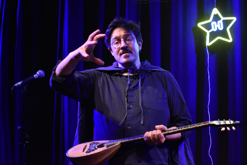 A man in glasses holds a stringed instrument and gestures in front of a purple curtain