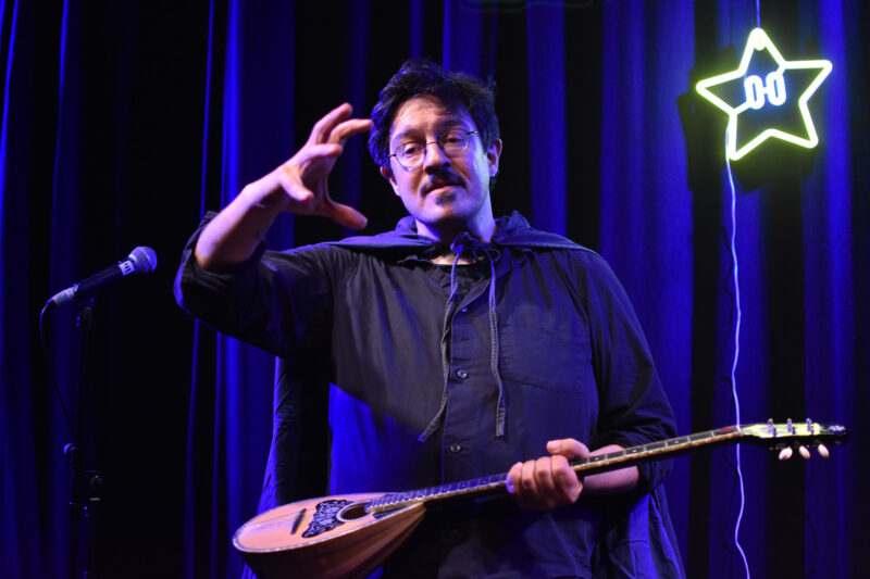 A man in glasses holds a stringed instrument and gestures in front of a purple curtain