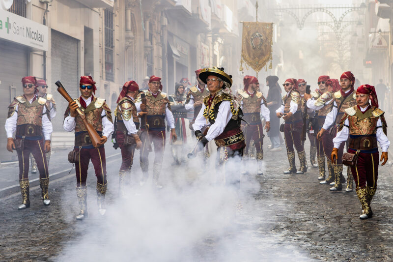 A line of men in gold and black costumes with red headscarves, some of them holding large old-fashioned guns (blunderbusses). The street is filled with smoke