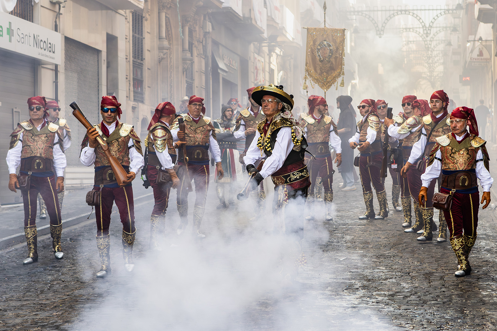 A line of men in gold and black costumes with red headscarves, some of them holding large old-fashioned guns (blunderbusses). The street is filled with smoke