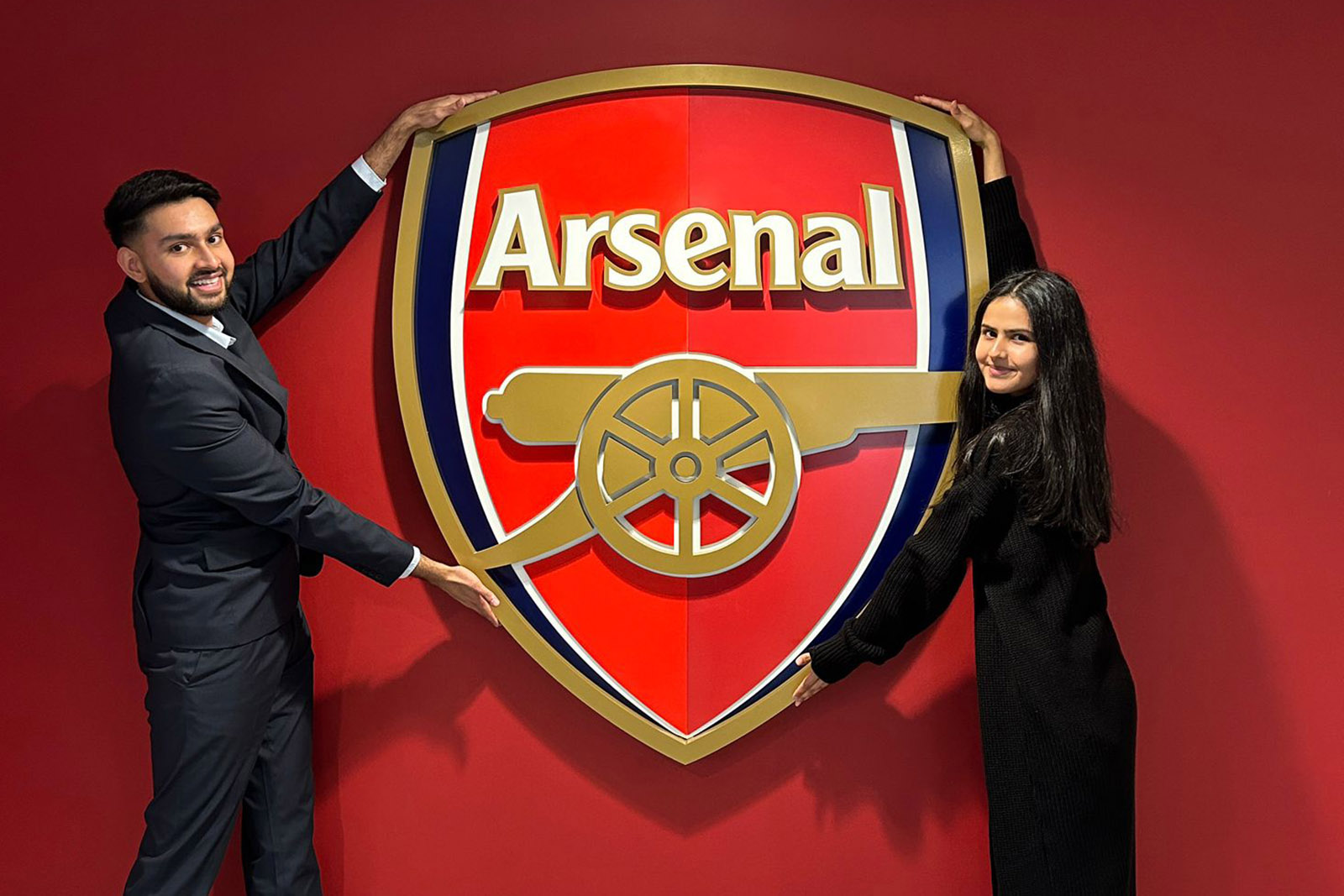Two people smile and pose with a huge Arsenal crest against a red wall