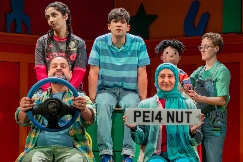 A production still of the Planet Omar cast (clockwise from bottom left): Umar Butt (dad, holding steering wheel), Emaan Durrani (Maryam), Justin Kendal-Sadiq (Omar), Izzy Coward with Esa puppet, Aizah Khan (mum, holding number plate)