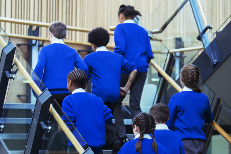 A stock image of a rear view of group of pupils wearing blue school uniforms walking up stairs in school