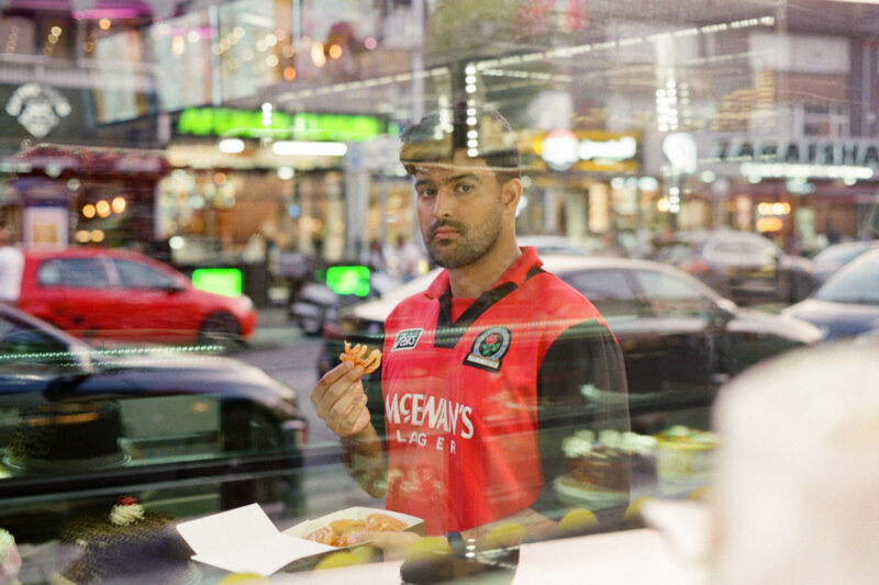 A photograph of Sufiyaan Salam, wearing a vintage Blackburn Rovers shirt with McEwan's Lager branding on the front and holding a container of street food on the Curry Mile, taken through the window of a shop