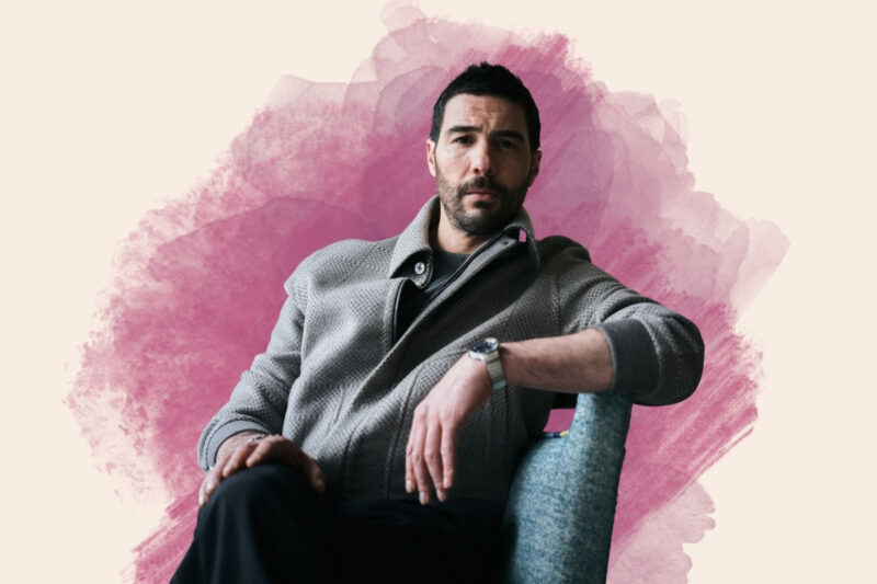 A composite image of French-Algerian actor Tahar Rahim, with a portrait photograph of him seated cross-legged, wearing a grey jacket, superimposed on a white and red background