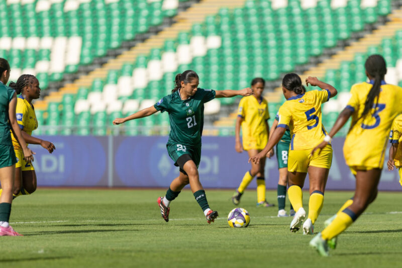 A woman in green with a football at her feet, while players in yellow cluster around her. In the background are the green seats of a stadium.