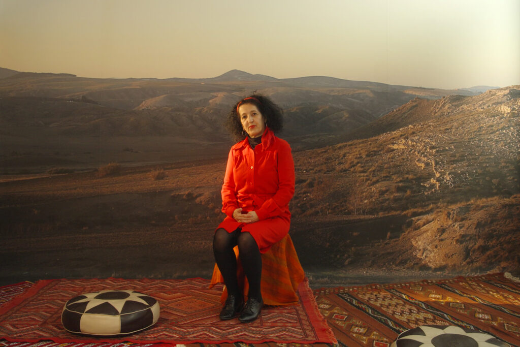 A photograph of French-Algerian artist Zineb Sedira, wearing a red dress and seated with her hands clasped together on her lap in front of a large image of an arid, hilly landscape