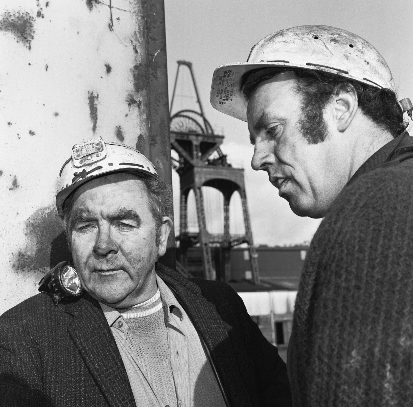 A black and white photograph featuring miners' union leader Joe Gormley (on the left), and another man at a coal mine. Both are wearing miners' helmets and appear to have coal dust on their faces, with pit head winding gear in the background.