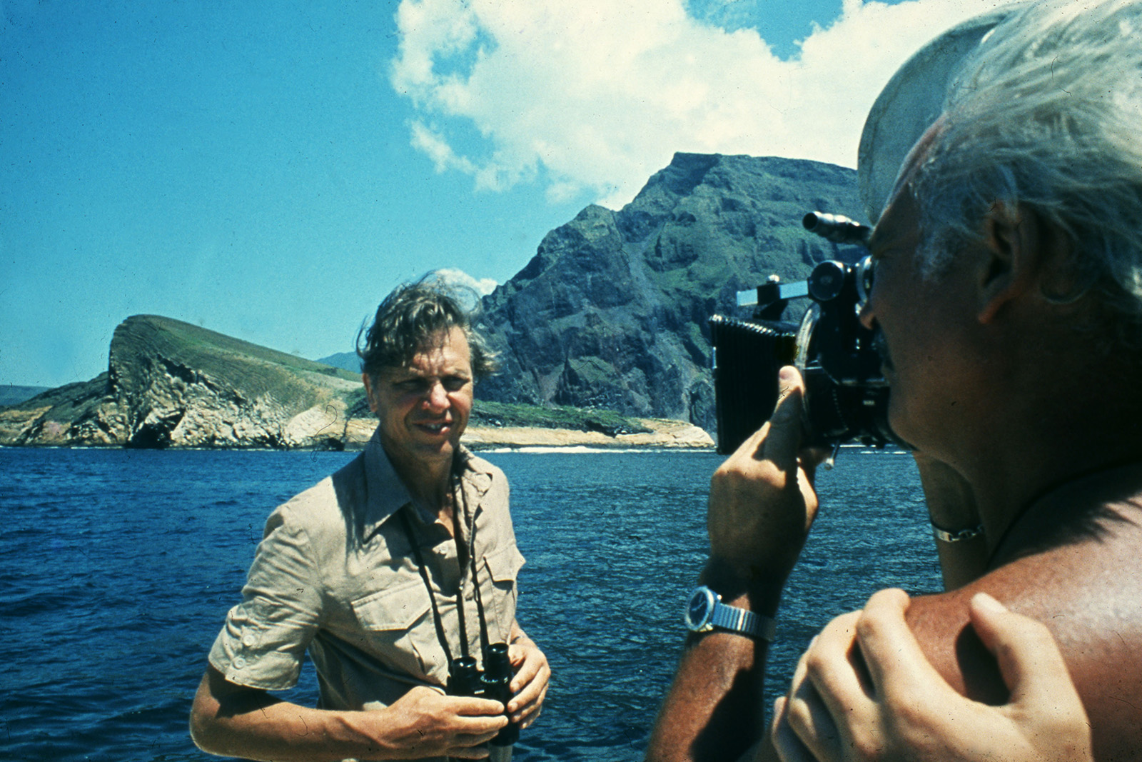 A photograph of David Attenborough during production of Life on Earth, standing on a boat with a rocky shoreline in the background. Wearing a khaki shirt and with hands resting on a pair of binoculars slung around his neck, he's being filmed by a shirtless cameraman, shown in the foreground of the image
