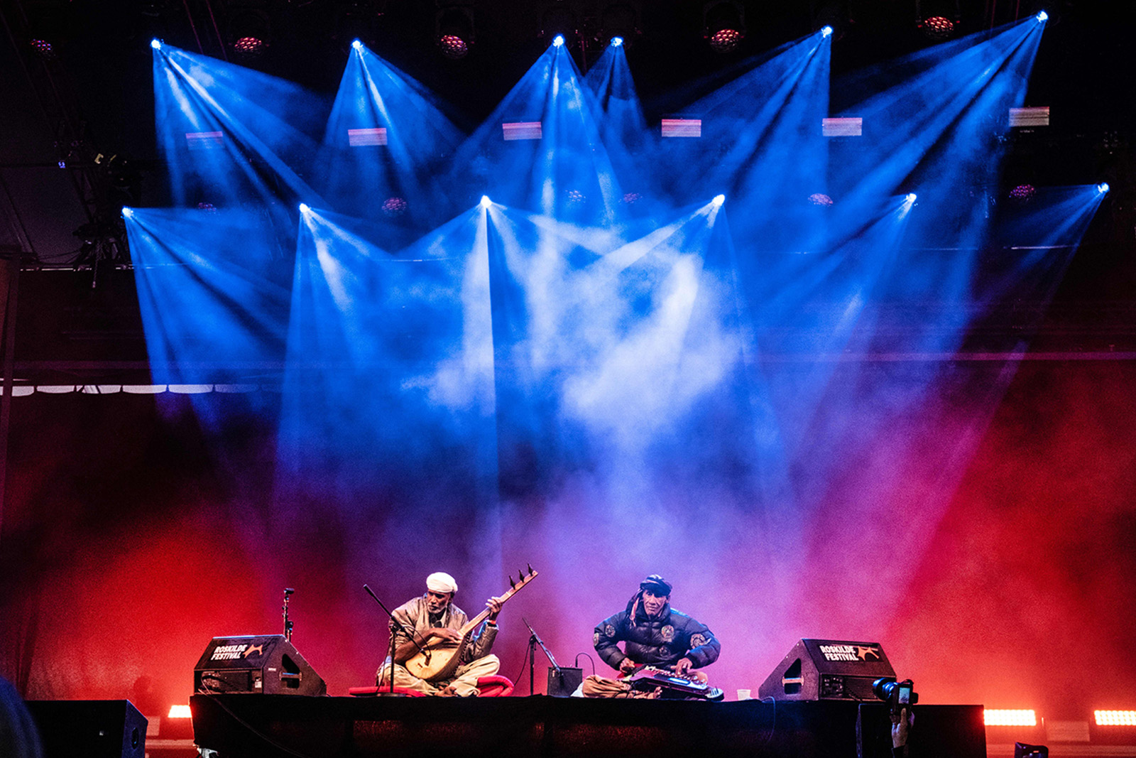 A photograph of Ustad Noor Bakhsh, seated with his benju instrument nestled in his crossed legs beside another musician, performing on stage at the Roskilde music festival in 2023