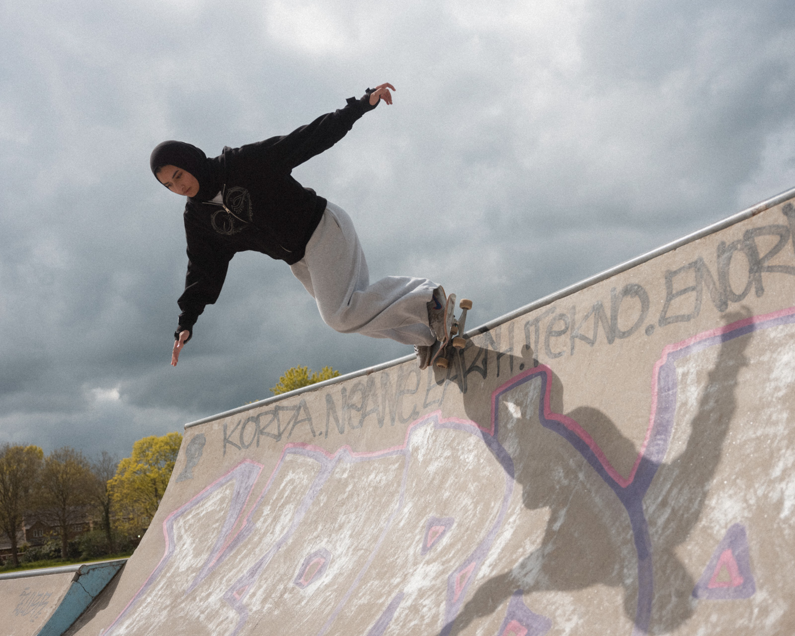 A photograph of Huda Kherati in action, captured balanced on the lip of a half pipe with her arms outstretched while skateboarding 