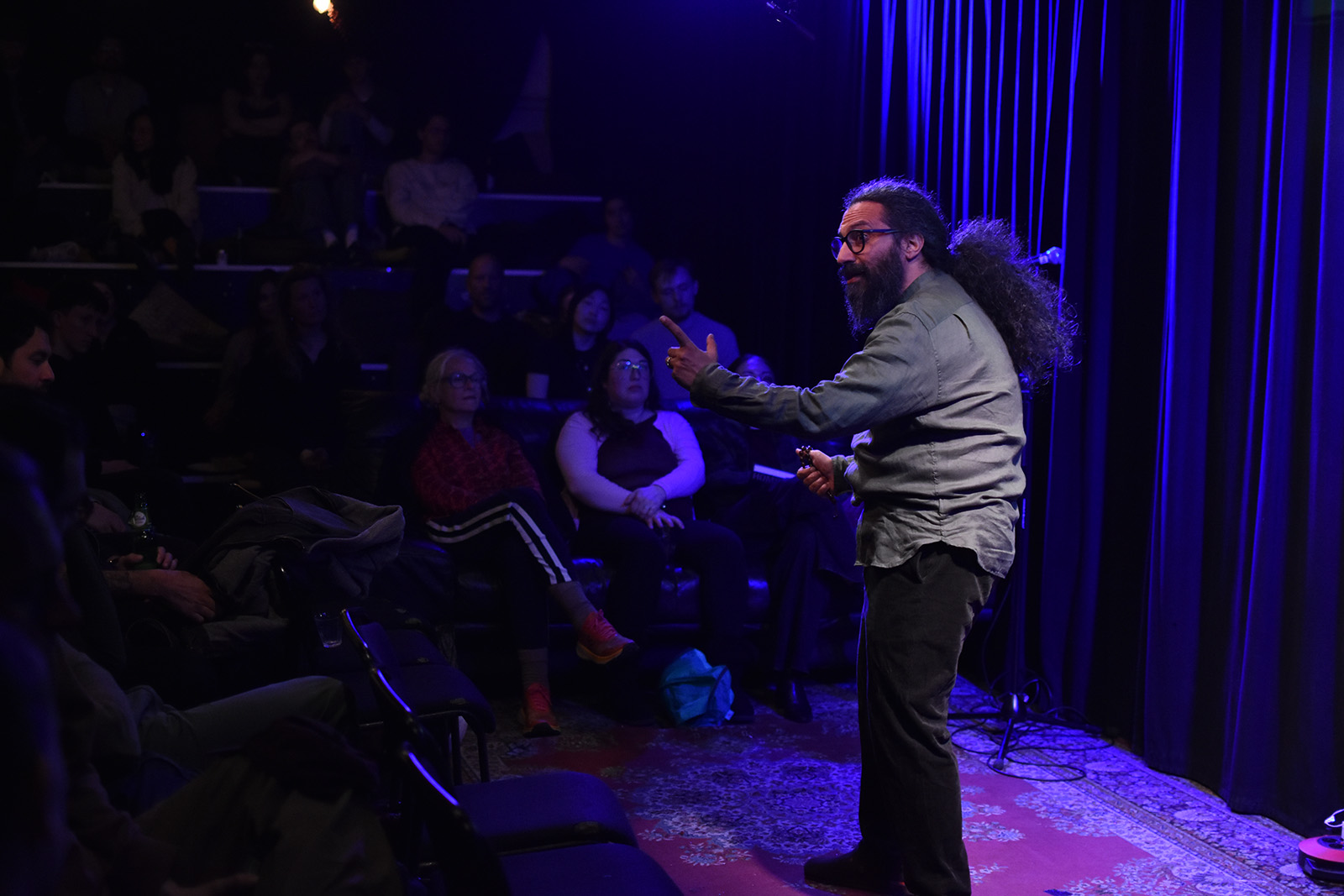 A man with glasses and long hair tied back gestures expressively with one arm, holding a mic in the other, with a dimly lit audience surrounding him close by.