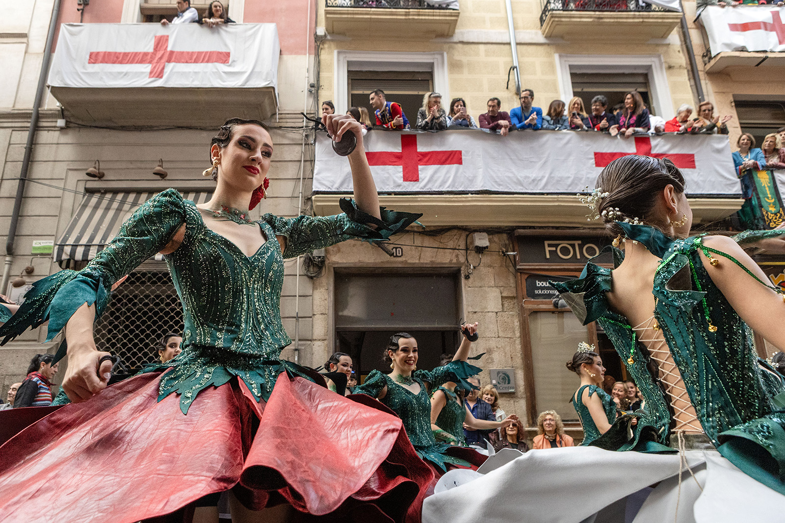 Dancers in green tops detailed with gold thread and red or white skirts, with their left arms aloft. Behind them, spectators watch from a balcony with St George's crosses displayed beneath it.