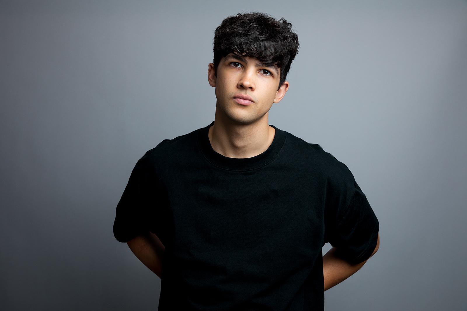 A portrait photograph of Planet Omar lead actor Justin Kendal-Sadiq, standing with his arms behind his back wearing a black T-shirt