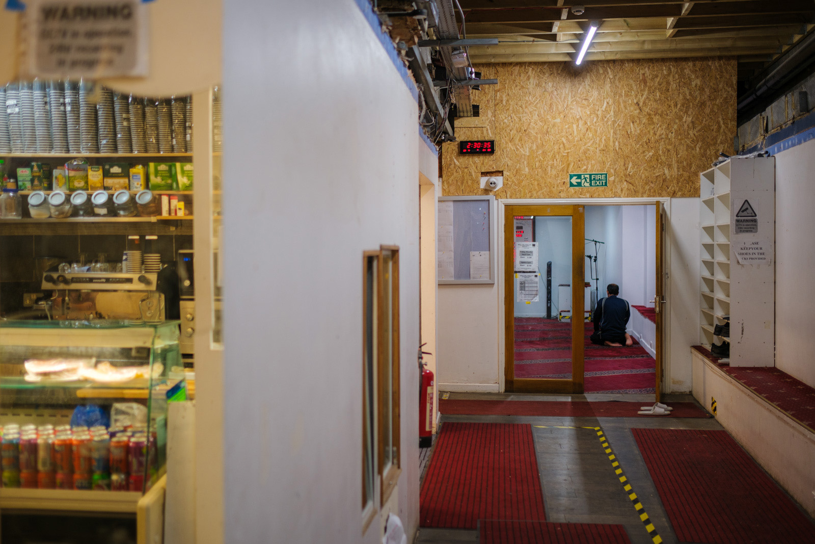A photograph of the entrance to the small mosque tucked away  down a corridor at the back of Queensway Market. Inside a man with his back to the camera is kneeling down to pray, while to the right of the doorway shoes are placed on shelving