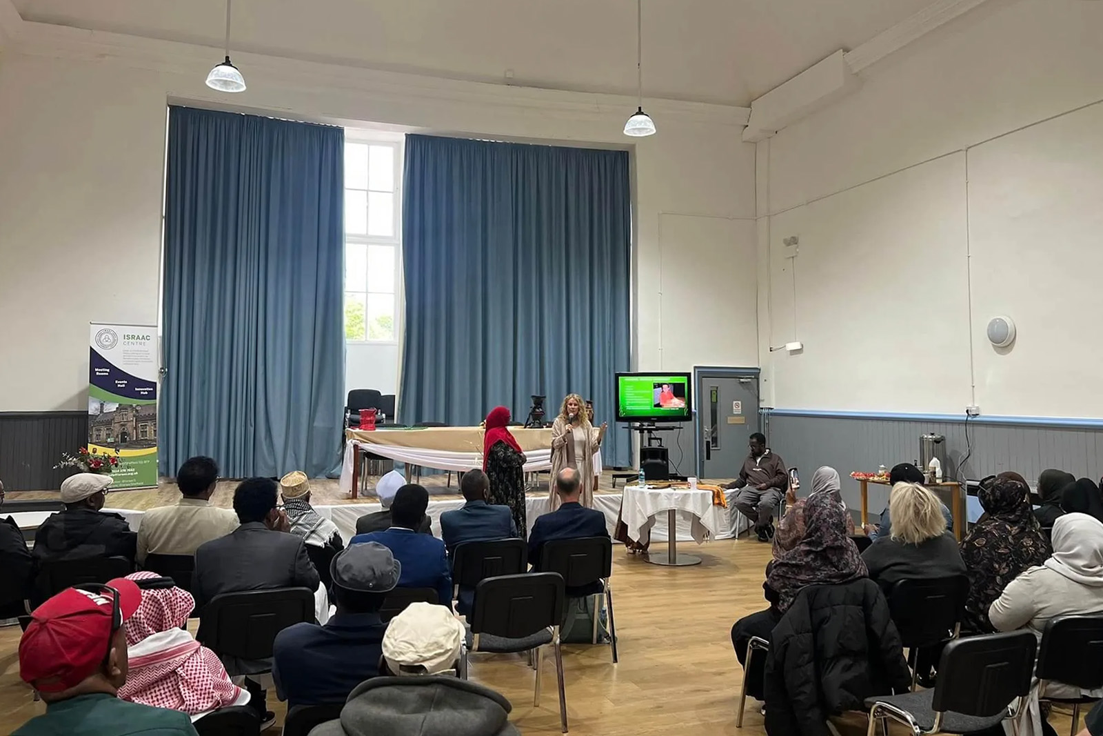 A photograph taken inside the Israac Somali Community Association, showing a woman with a microphone addressing a seated crowd of people in a large room with a high ceiling
