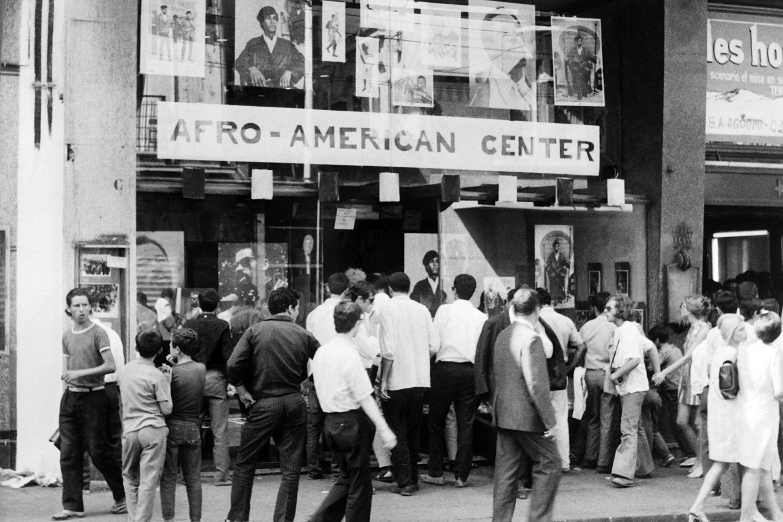 A black and white archive photo of visitors outside the Afro-American Center in Algiers, July 1969, during the opening parade for the first Pan-African Cultural Festival. Large portrait photos of Black Panther leaders including Huey Newton and Eldridge Cleaver are displayed in the center's windows