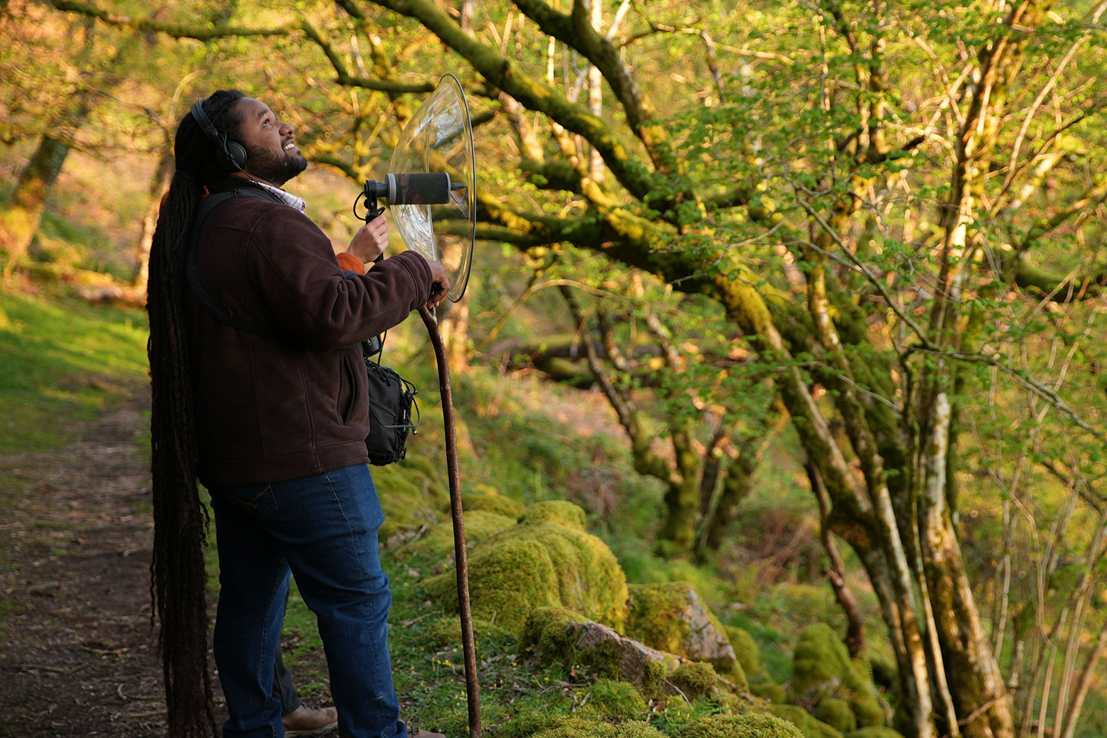 A photograph of natural history broadcaster Hamza Yassin, wearing headphones attached to a microphone held by a colleague, listening to birdsong at dawn, in an ancient woodland on Dartmoor