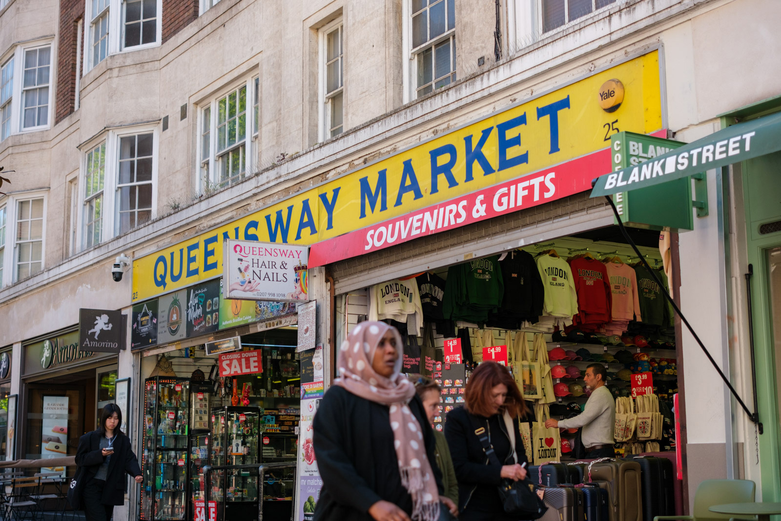 A photograph of the blue on yellow 'Queensway Market' signage at the entrance to the market on a busy street with shoppers walking by