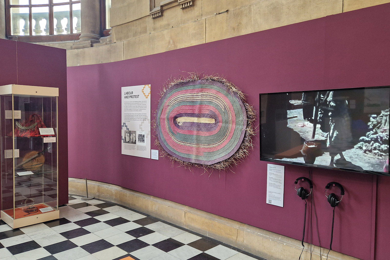 A photograph of a section of the Cartwright Hall Art Gallery exhibition with an information board, titled Labour and Protest, and a Somali rug. The exhibition highlights how Somali Village members protested about their pay and living conditions