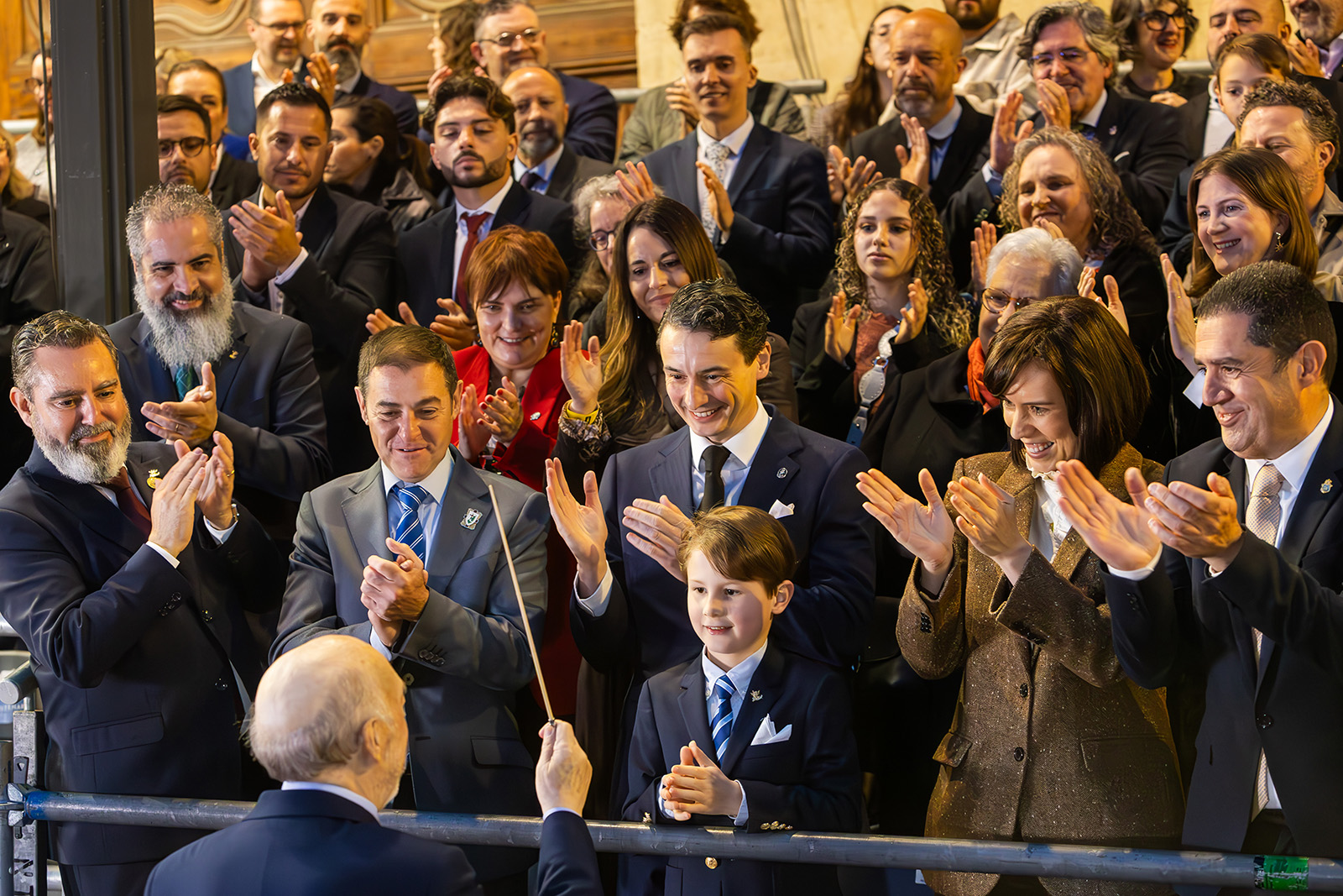 A group of smartly dressed clapping people as a young boy in a suit faces a conductor holding a baton.