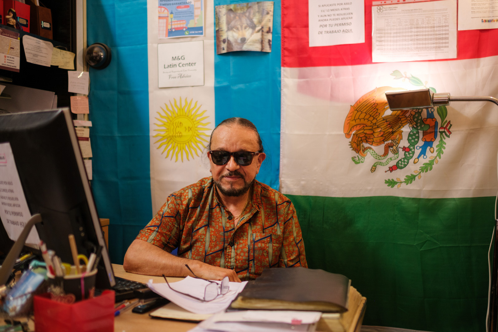 A photograph of lawyer George Cubillos, wearing sunglasses and sitting at his desk in his unit in Queensway Market, from where he has run an advice service for Latin American immigrants for many years