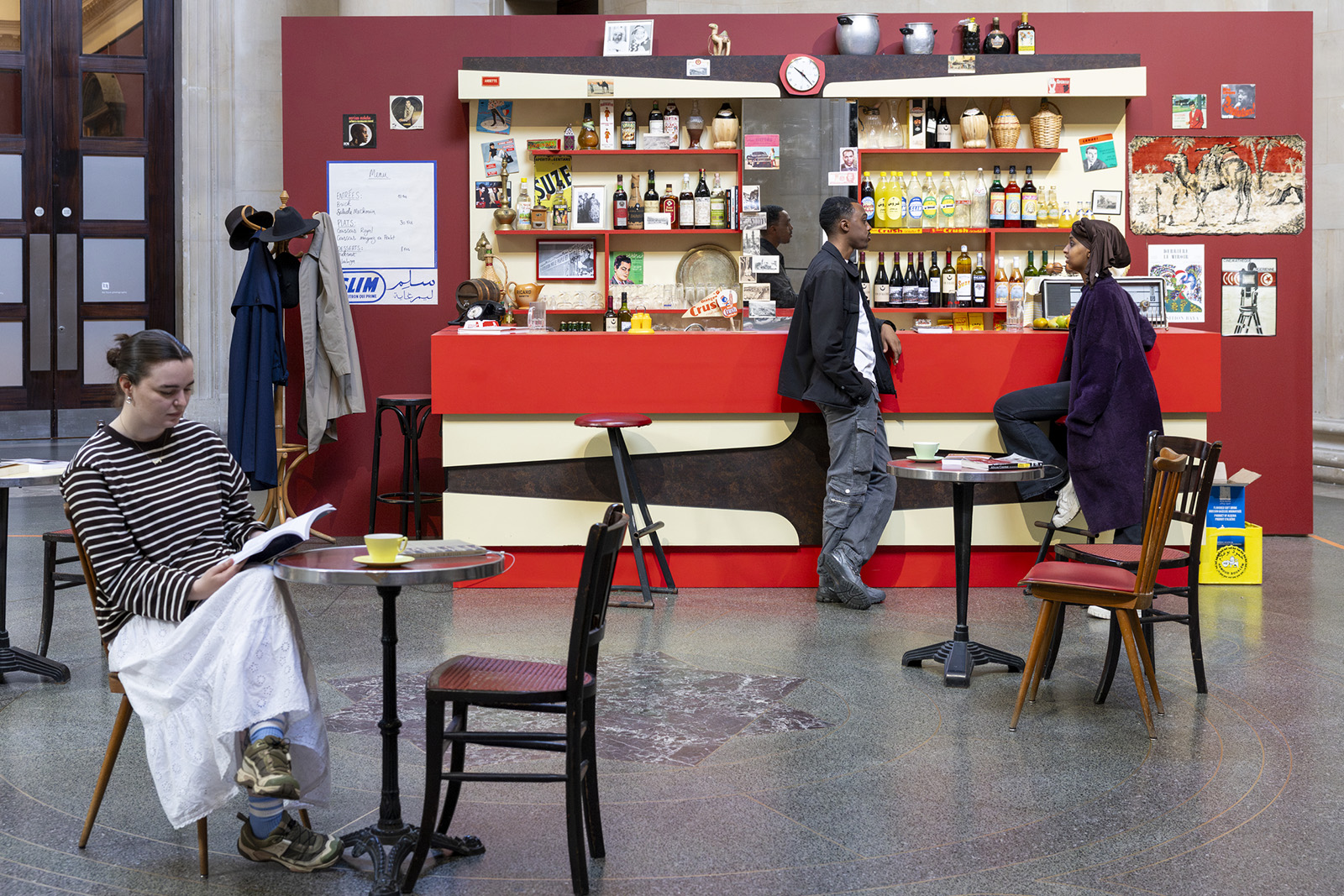 A photograph of part of Zineb Sedira's installation for her 2026 Tate Britain commission, featuring a recreation of a 60s Paris cafe, with two people leaning against the bar in conversation and a woman seated at a table in the foreground reading a book 