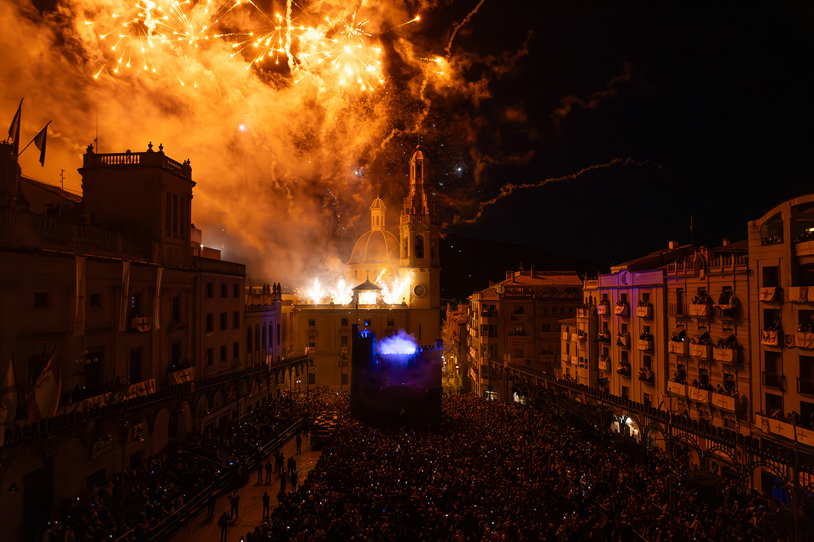 A wide-angle shot of an orange and yellow fireworks display, looking almost like a ball of fire in the sky, illuminating a square of grand buildings, including a domed building in the centre. The lights of people's phone cameras are just visible in the darkness below.