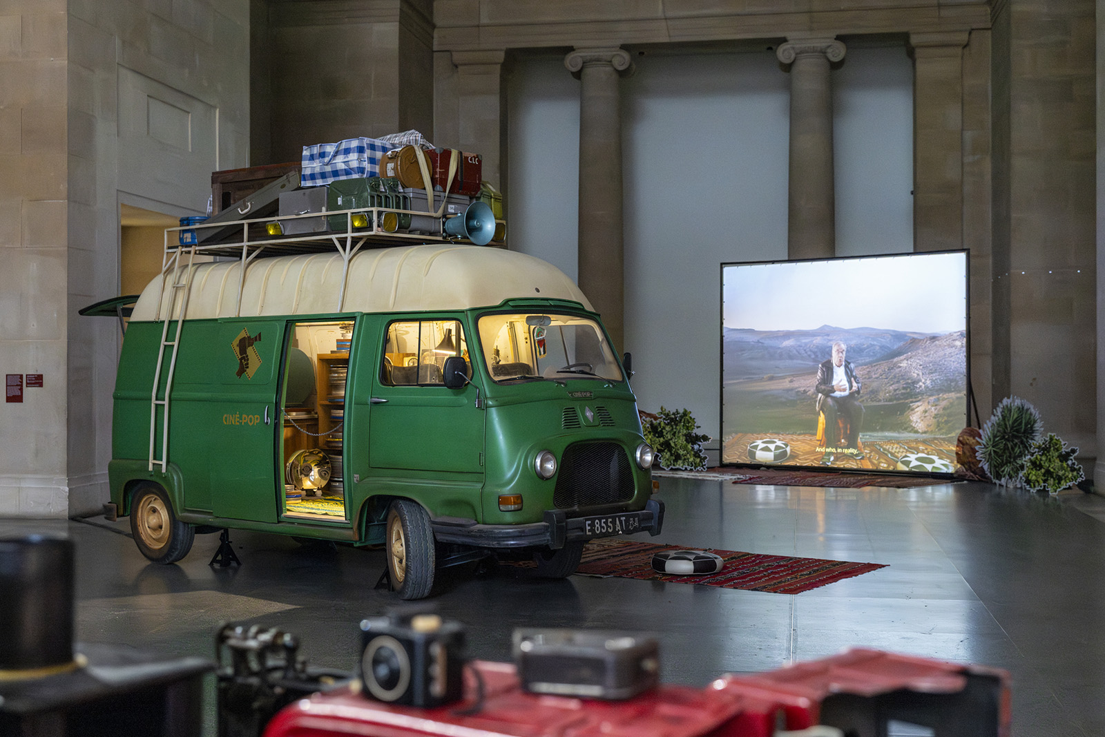 A photograph of Zineb Sedira's recreation of a Ciné-Pop mobile cinema van for her 2026 Tate Britain commission installation. The green van sits beside a large screen projecting Sedira's interview with Algerian film critic and historian Ahmed Bedjaoi