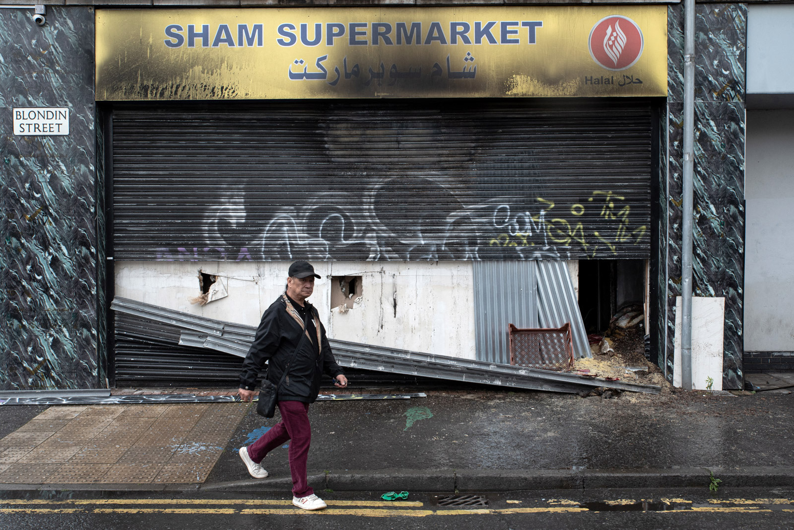 A photograph of a man walking past a burned out shop on Blondin Street, Belfast, which was attacked during the disorder on the night of 9 June 2026
