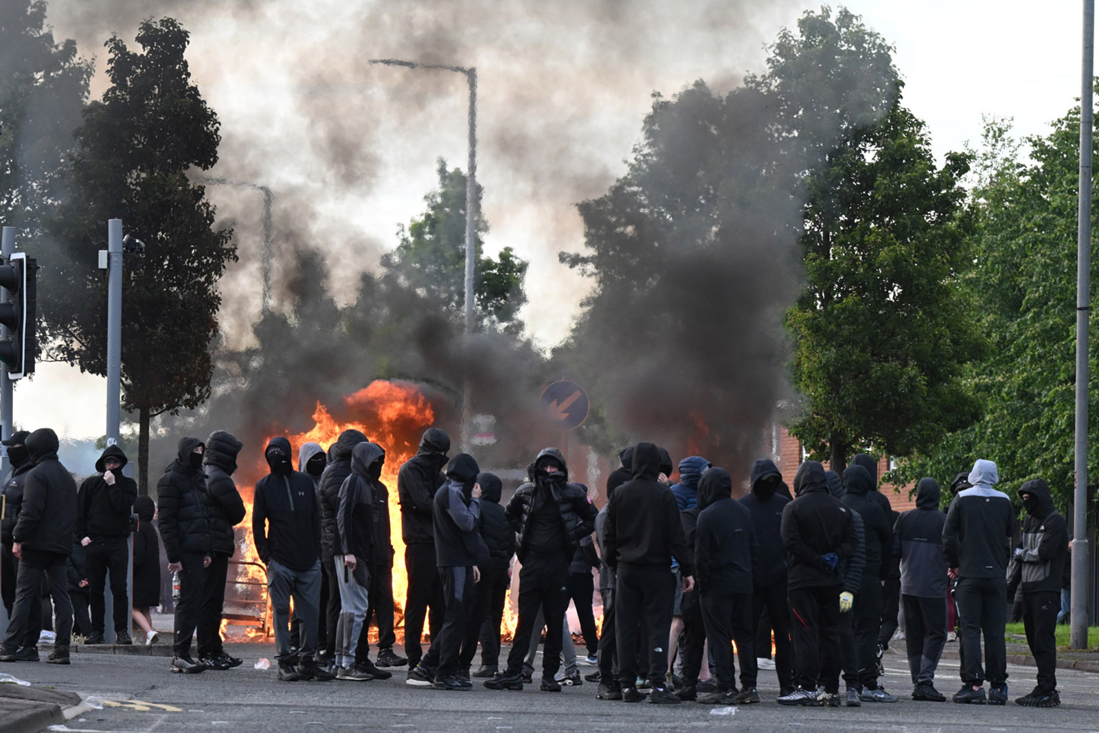 A line of masked people in black with a fire burning behind them and smoke in the air