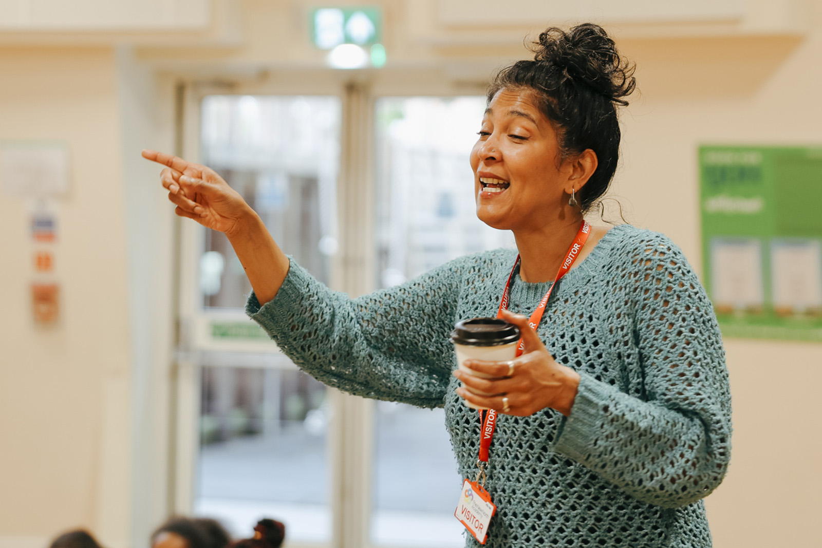 A photograph of Khiyo singer Sohini Alam, cup of coffee in hand and visitor lanyard round her neck, during a music workshop in a classroom at New North Academy primary school in Islington, London