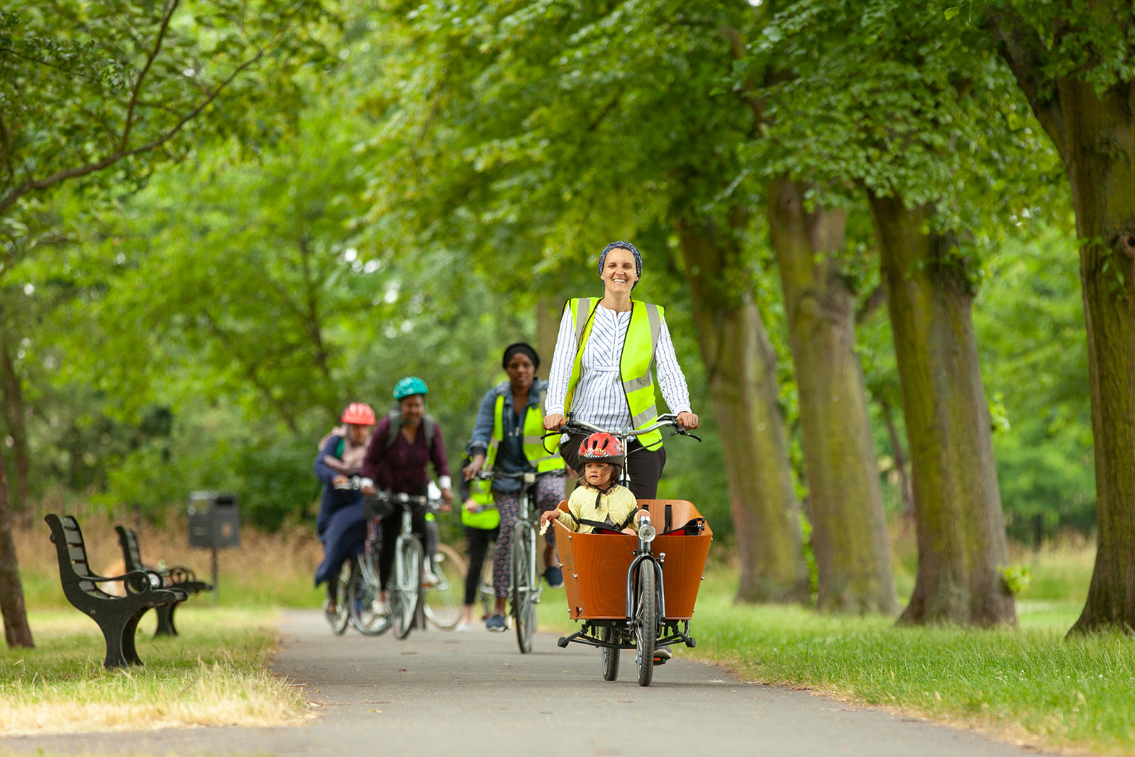 A photograph of a Cycle Sisters event, featuring a group of women riding on a path in a park, led by a smiling mother with her child on a cargo bike