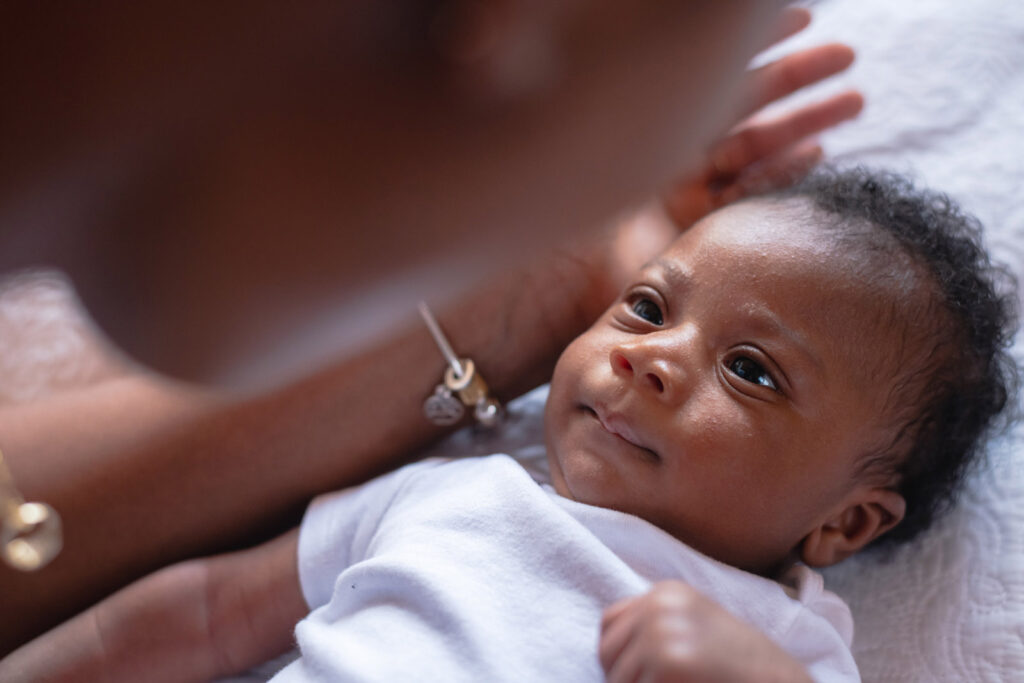 A stock image of a young black mother is leaning over her newborn son, who is wearing a white jumpsuit and staring at her intently with a small smile on his face