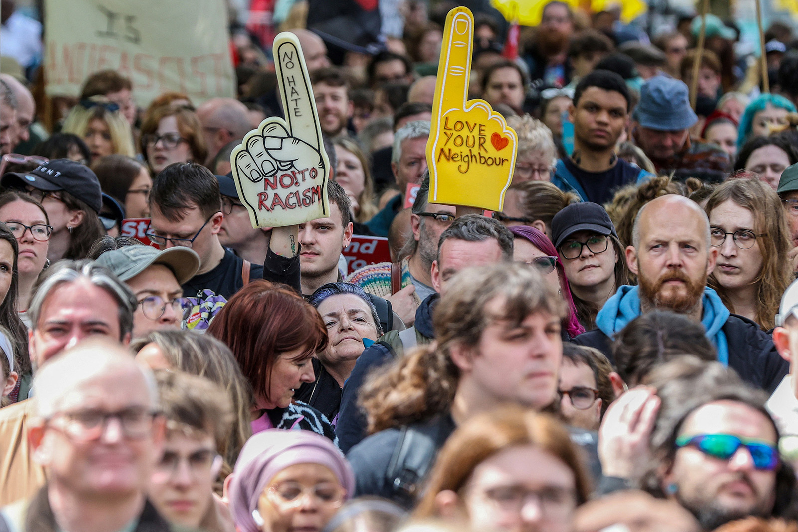 A photograph of a crowd of people at an anti-racism demonstration in Belfast on 13 June 2026, following rioting in the city, with foam hands visible with 'No hate, No! To racism' and 'Love your neighbour' slogans