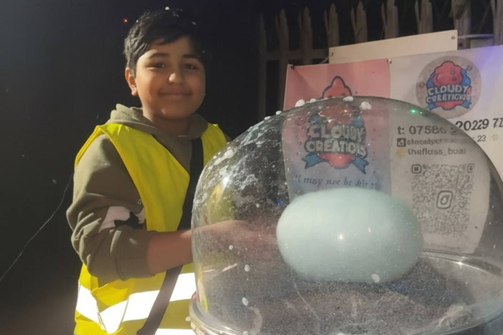 Hashir Aziz making candyfloss with a blob of sugar inside a sort of glass orb at the top of a machine. He's a young boy wearing a high vis top and is smiling.