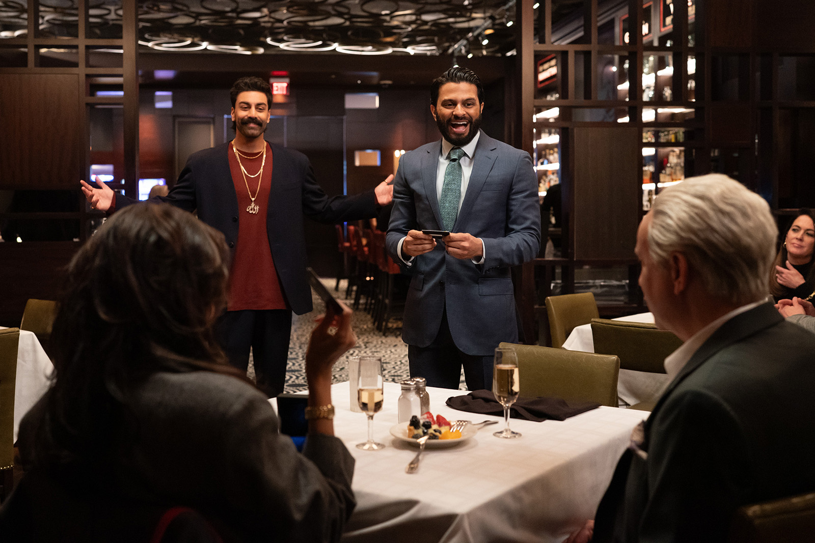 A photograph of Saagar Shaikh (left) and Asif Ali as brothers Raj and Mir Dar in Deli Boys, in a restaurant scene also featuring Poorna Jagannathan and Fred Armisen, who are seated at a table with their backs to the camera