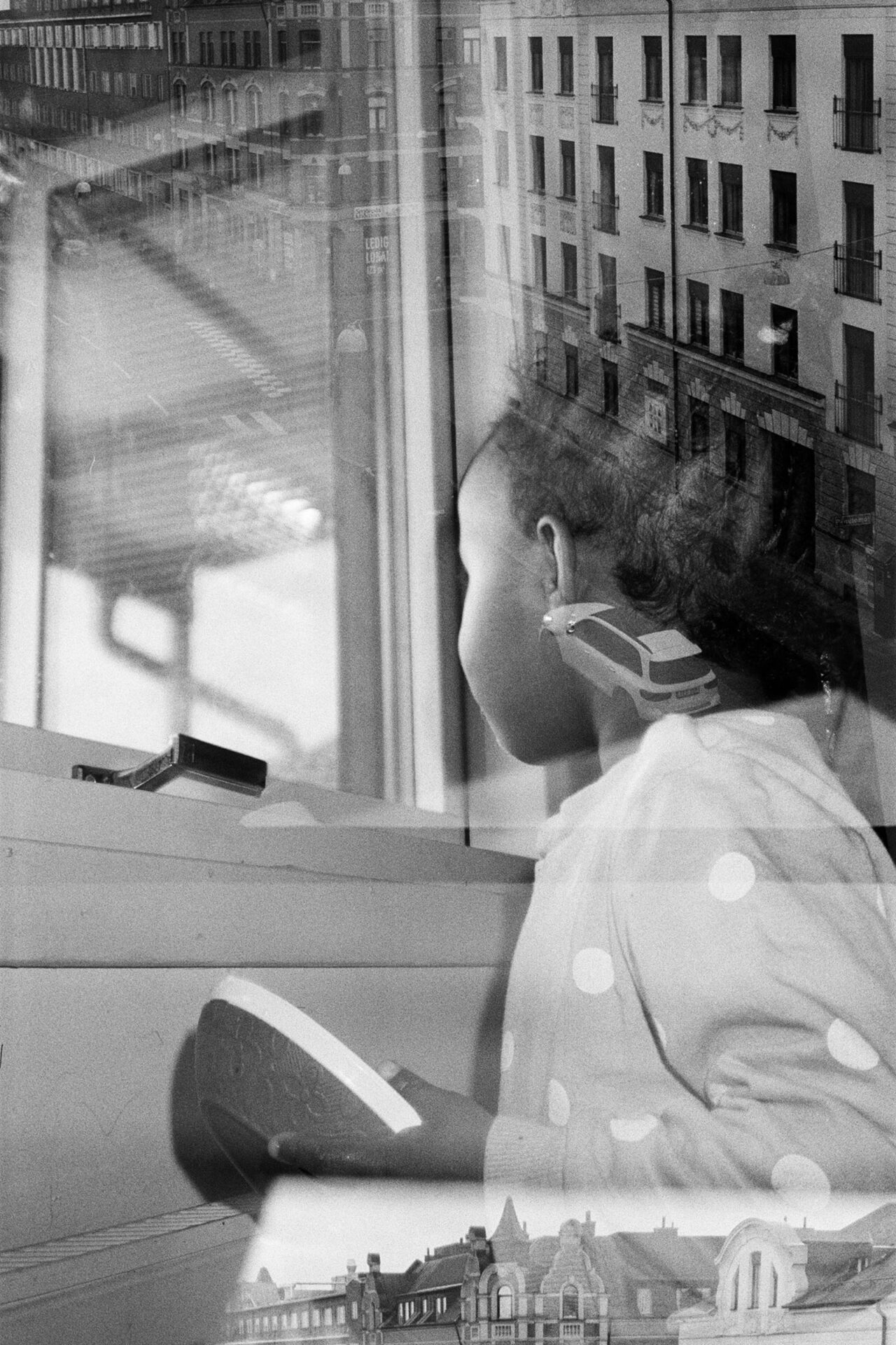 Photographer Ikram Abdulkadir's black and white double exposure image, with a street view featuring a car and buildings overlaid on a picture of a young girl looking out of a window