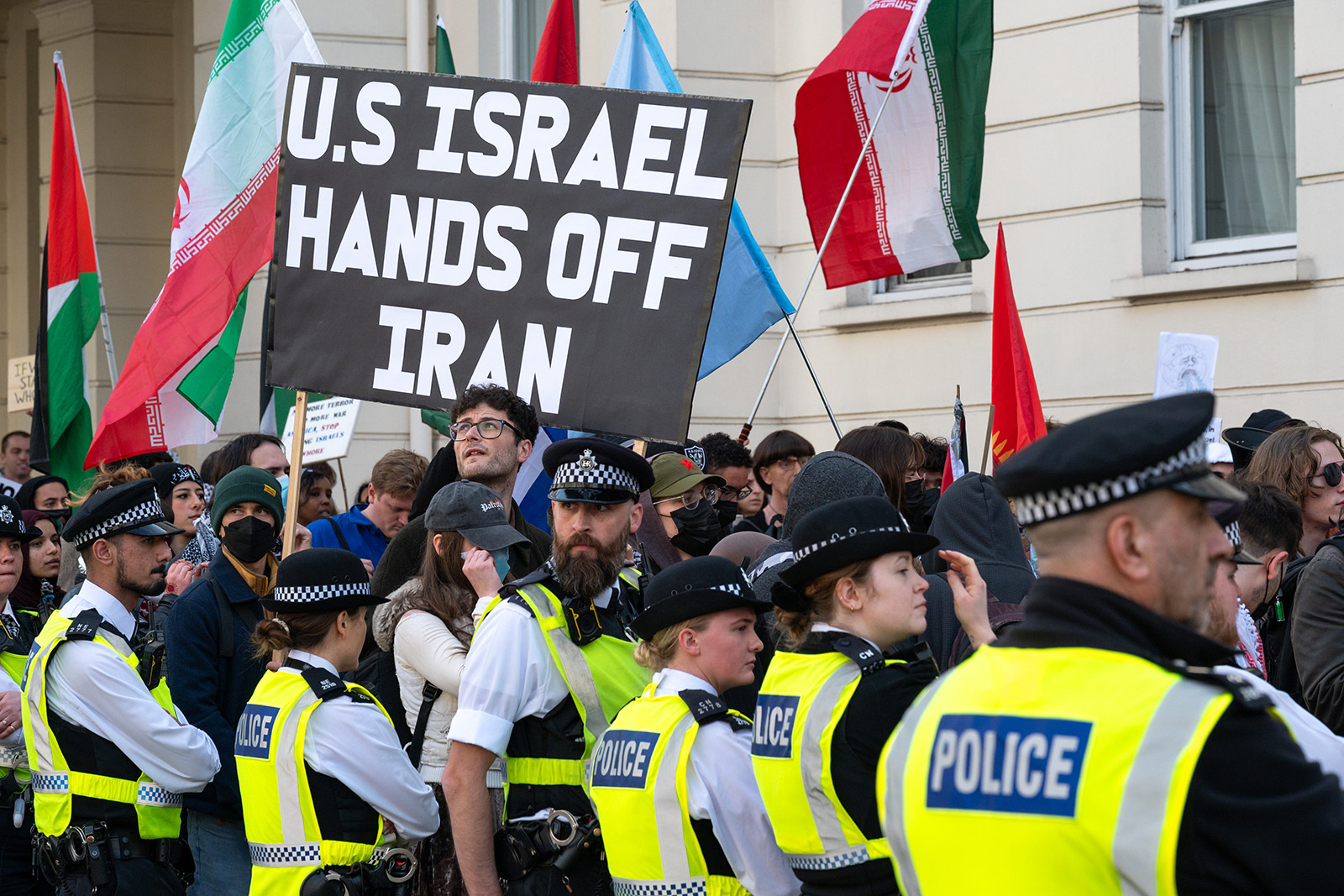 A photograph of a demonstration outside the US embassy in London in March 2026. In the foreground is a line of police officers, standing in front of protesters, one holding a a placard stating "US ISRAEL HAND OFF IRAN" and others waving Iranian flags