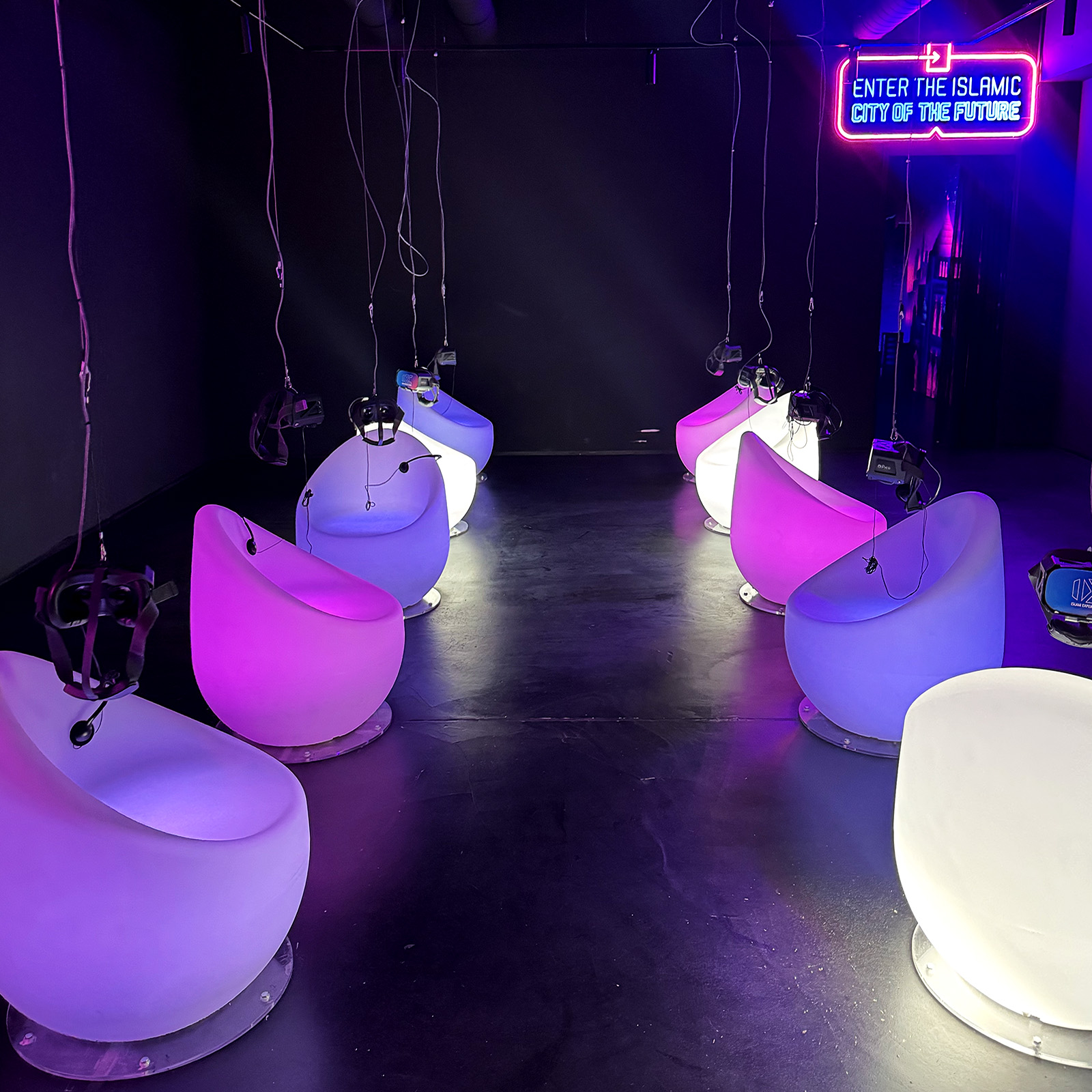 Plastic chairs lit pink and purple from within are arranged in two rows. A neon sign in one corner reads "enter the Islamic city of the future"