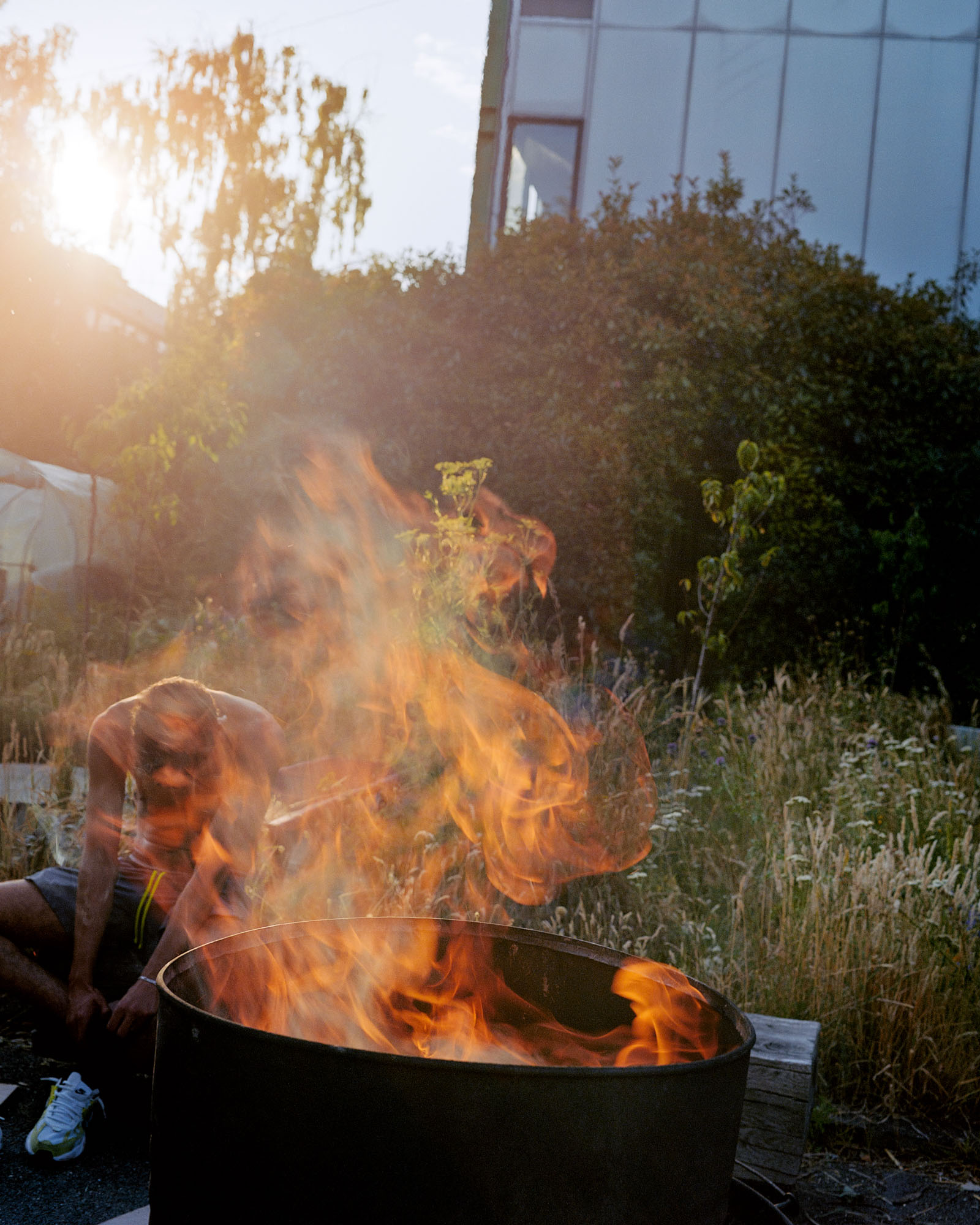 A photograph titled Petits Princes, featuring a seated young man wearing shorts and trainers but no shirt, with head bowed, viewed through the flames from a fire pit. From Camille Farrah Lenain’s monograph Made Of Smokeless Fire