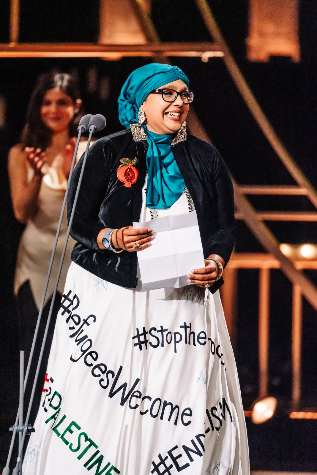 Onjali Q. Raúf at the 2026 Olivier Awards, where the play adapted from her novel The Boy at the Back of the Class won best family show. The author is wearing a white dress with slogans including #RefugeesWelcome